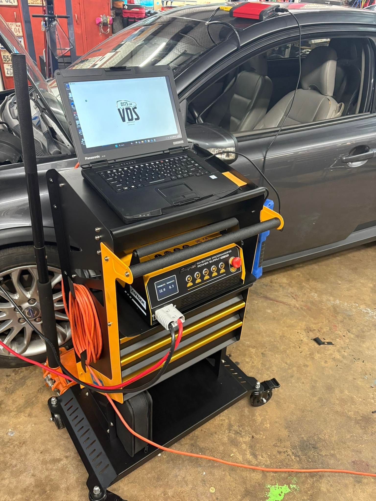 Laptop on a wheeled diagnostics cart beside a gray car in a garage, with cables and test equipment.
