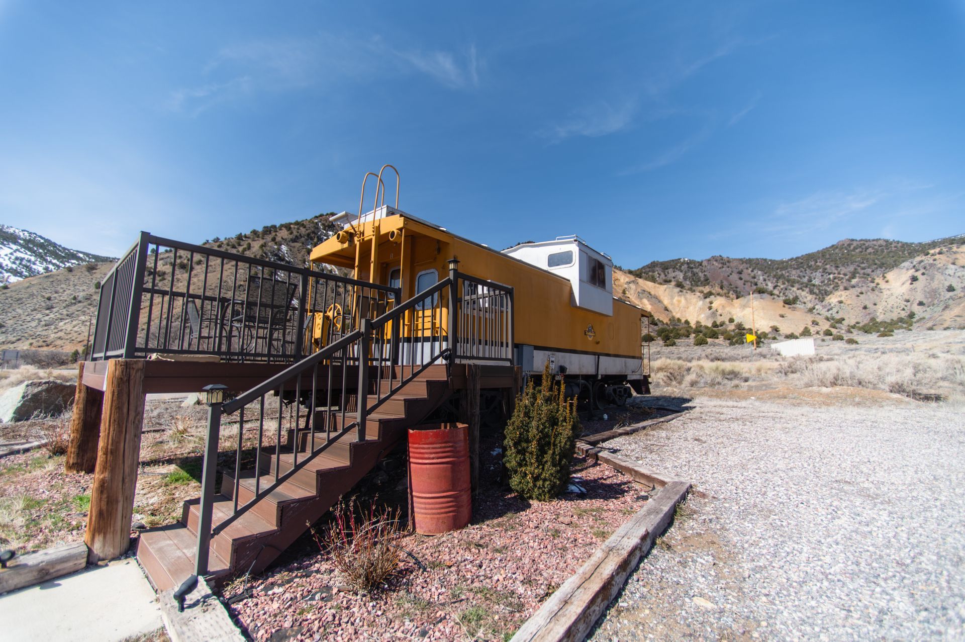 A yellow train car with stairs leading up to it is parked in a gravel lot.