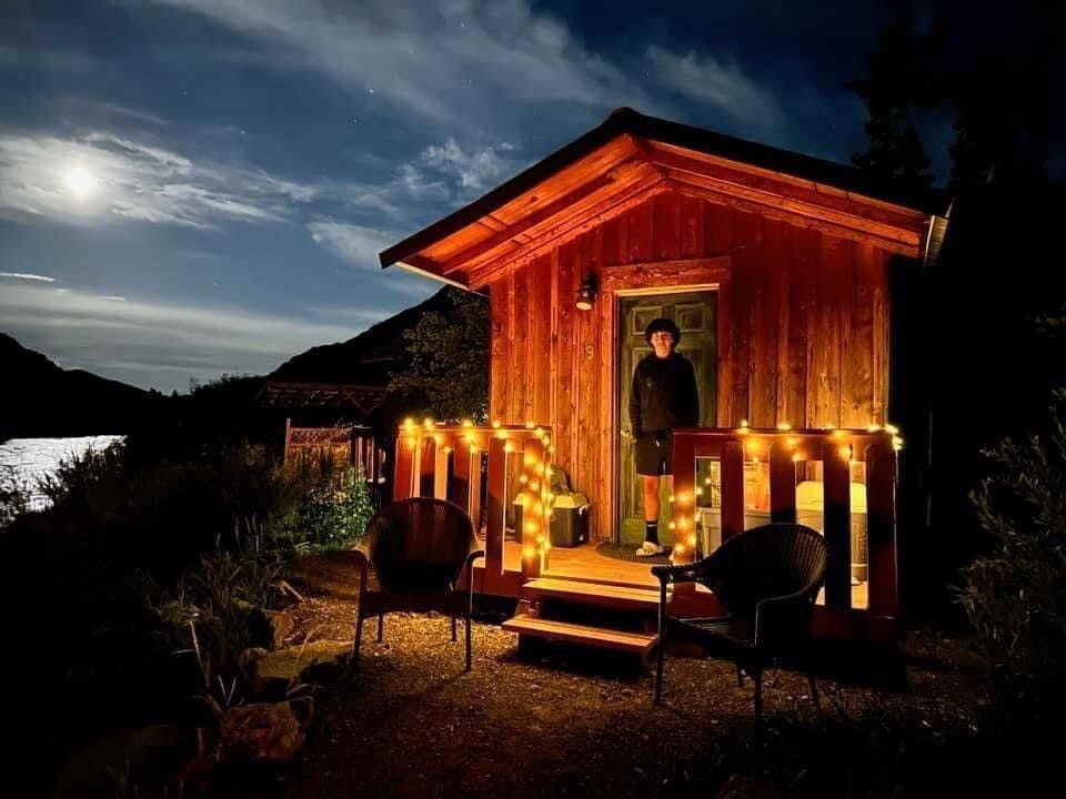 A man is standing in front of a small wooden cabin at night.