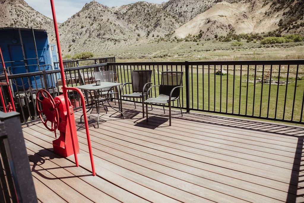 A wooden deck with chairs , tables and a railing with mountains in the background.