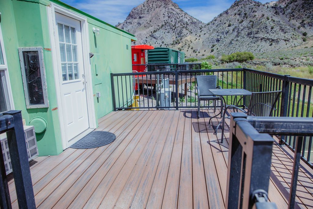 A wooden deck with a table and chairs in front of a green house with mountains in the background.