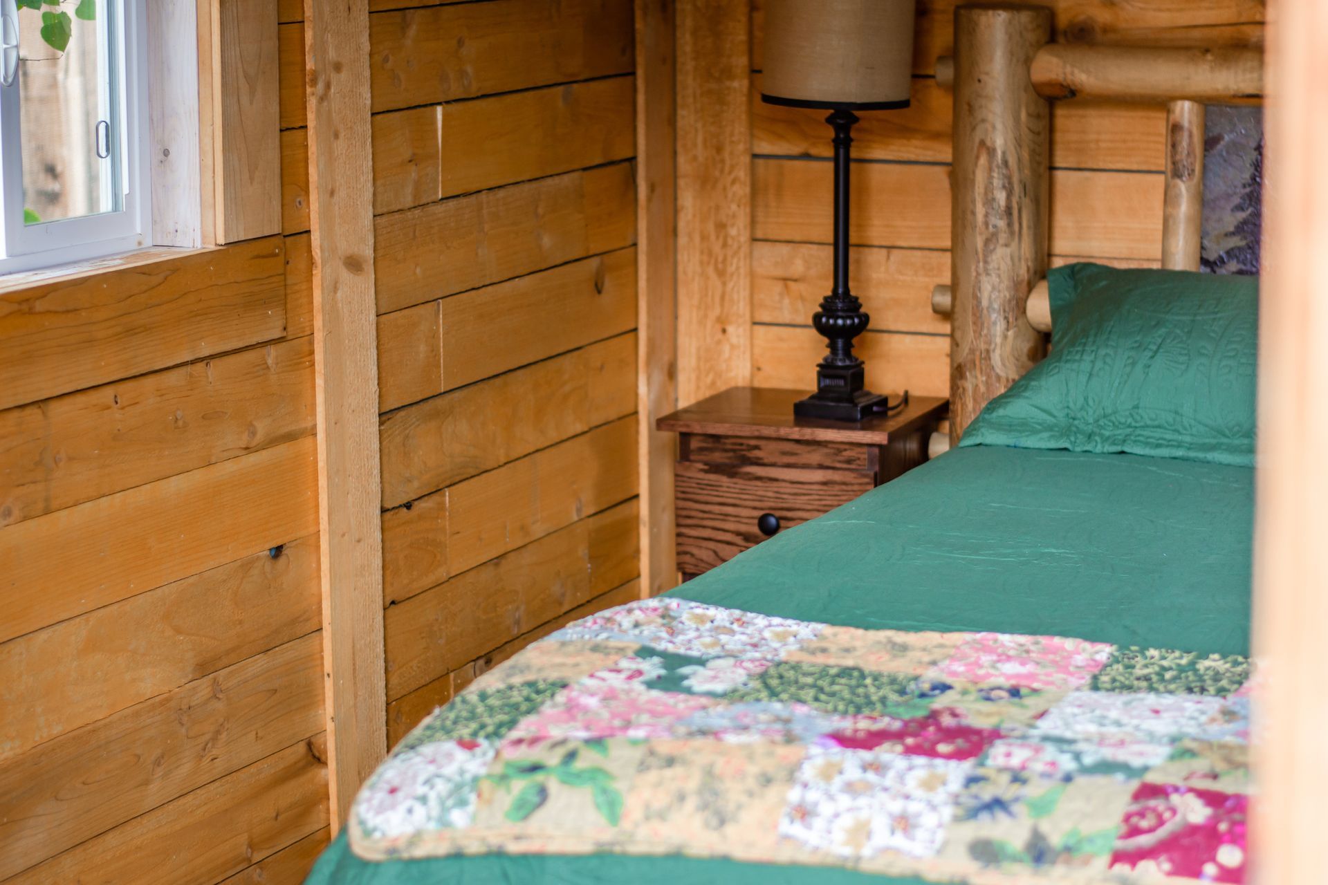 A bedroom in a log cabin with a bed , nightstand , lamp and window.