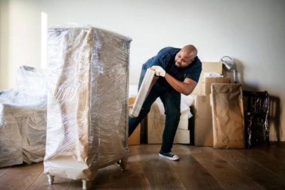 A man is carrying a piece of furniture in a room covered in boxes.