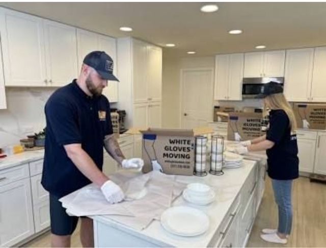 A man and a woman are packing plates in a kitchen with a box that says white gloves moving