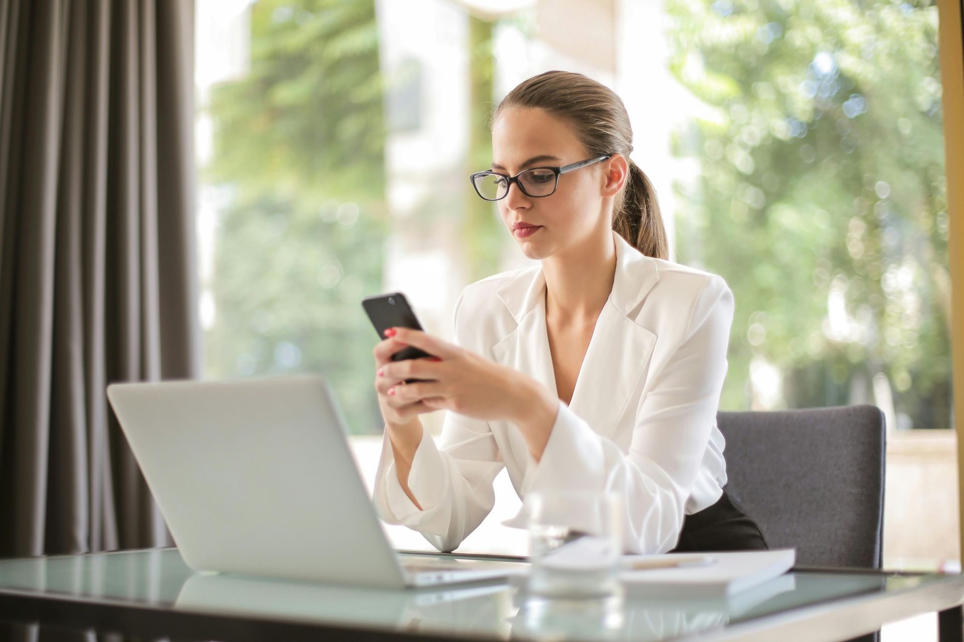 A woman is sitting at a desk using a laptop and a cell phone.