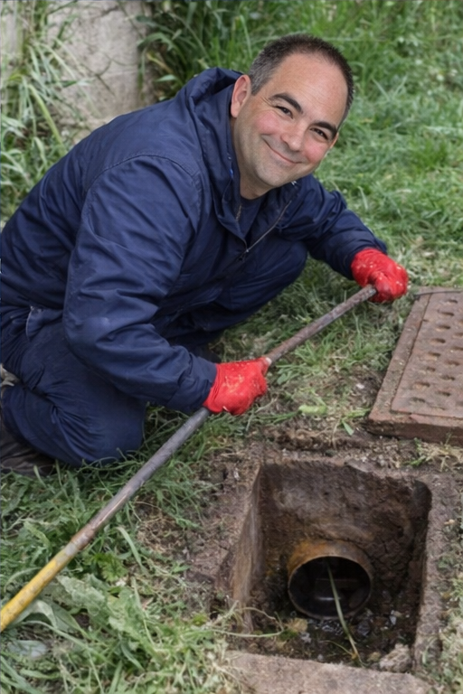 A person in a blue jacket and red gloves smiles while using a long tool to clear a pipe in a small outdoor excavation.