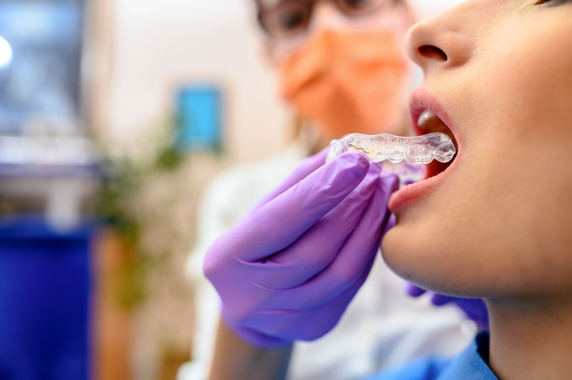 A dental professional, wearing a mask and gloves, places a clear retainer in a patient's mouth.
