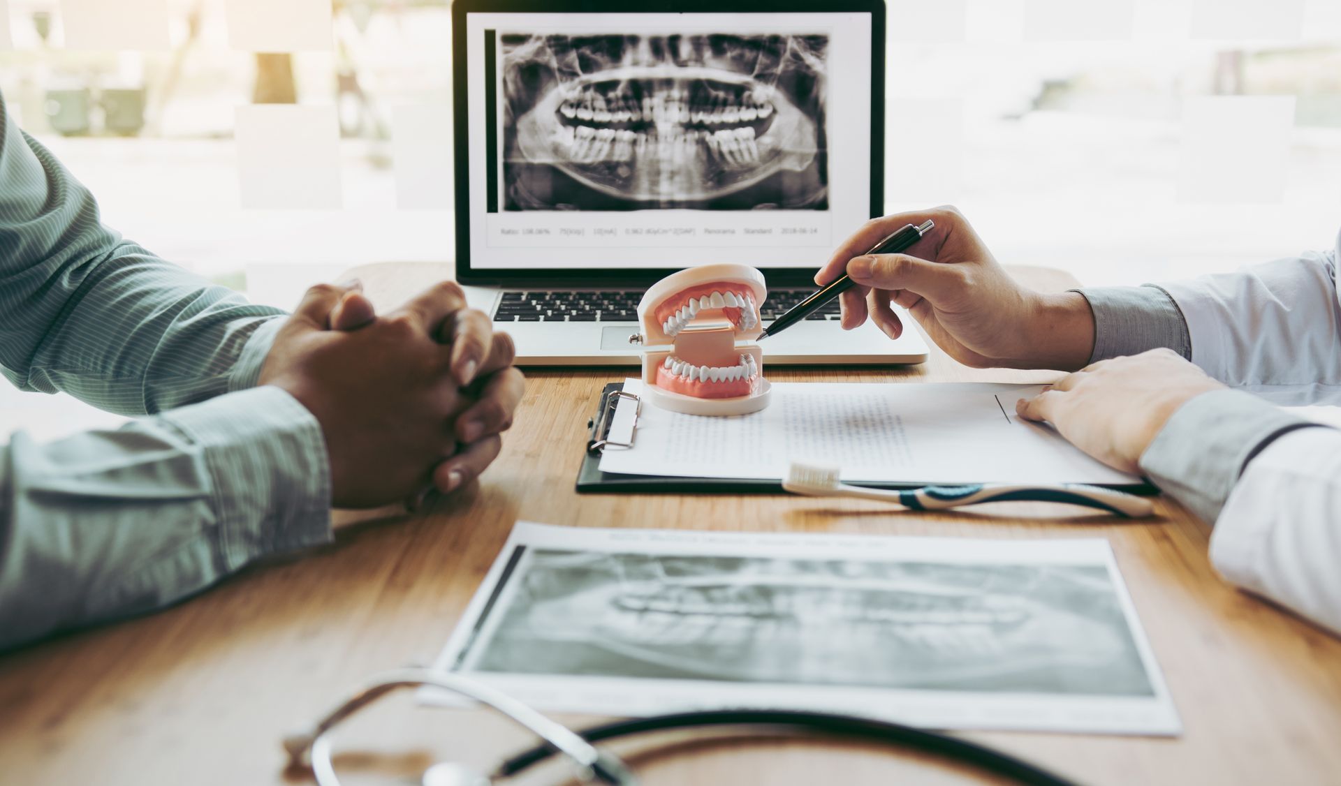 A dentist explains an x-ray with a model of teeth to a patient in a dental office, pointing with a pen.