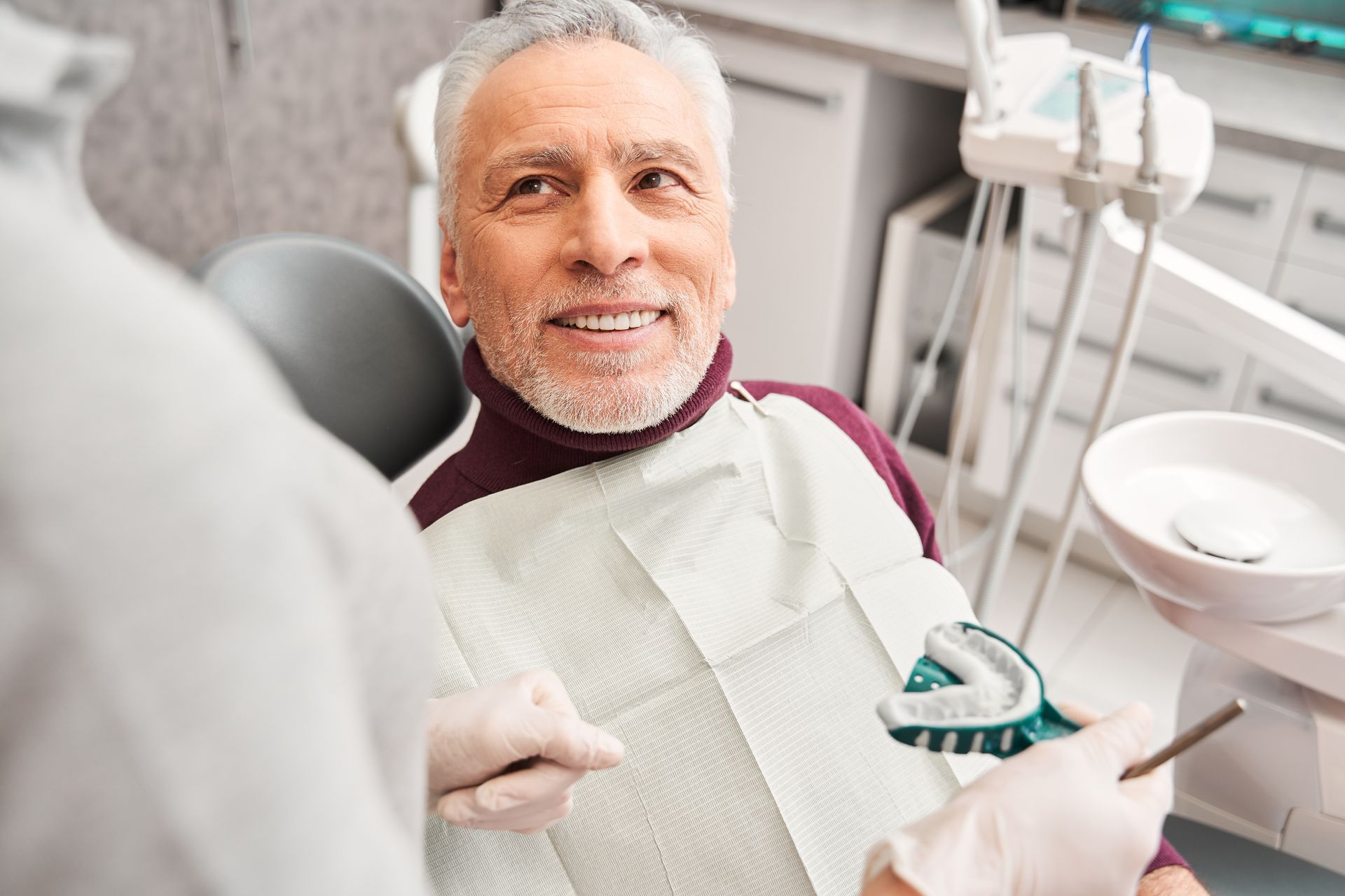 Smiling older man at a dentist's office, ready for a procedure. He's wearing a bib; a dentist holds tools.