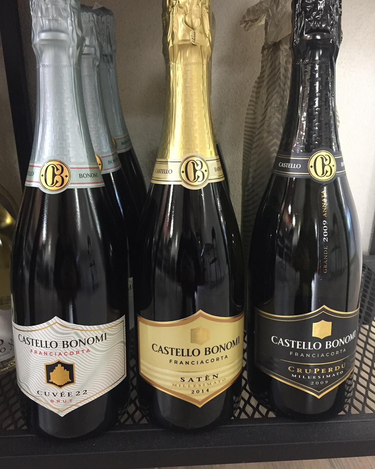 Tre bottiglie di spumante Castello Bonomi su uno scaffale, con etichette di vari colori.