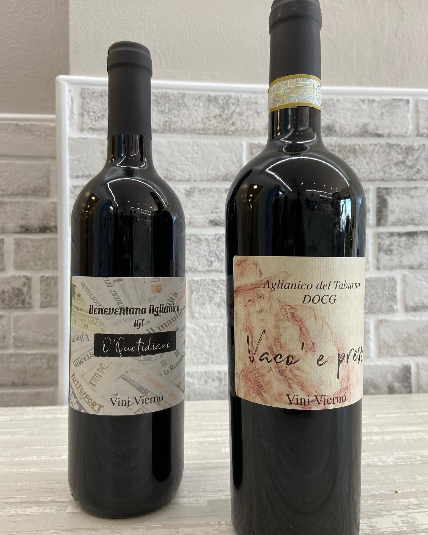 Due bottiglie di vino scuro su un tavolo con etichette, una alta e una più bassa, contro un muro di mattoni.