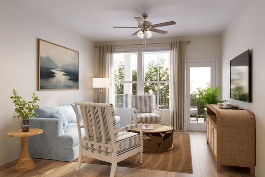 A sunlit living room with a blue sofa, patterned armchair, wood coffee table, TV console, and a coastal landscape painting.