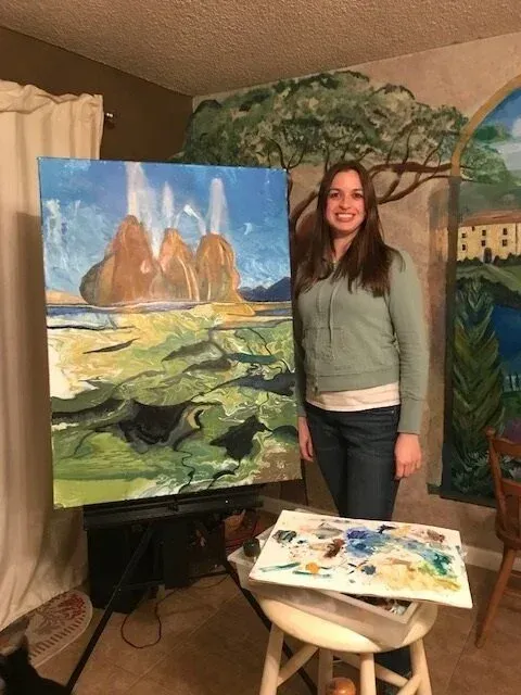 A person stands smiling beside a large painting of geysers and rocky terrain on an easel in a room.