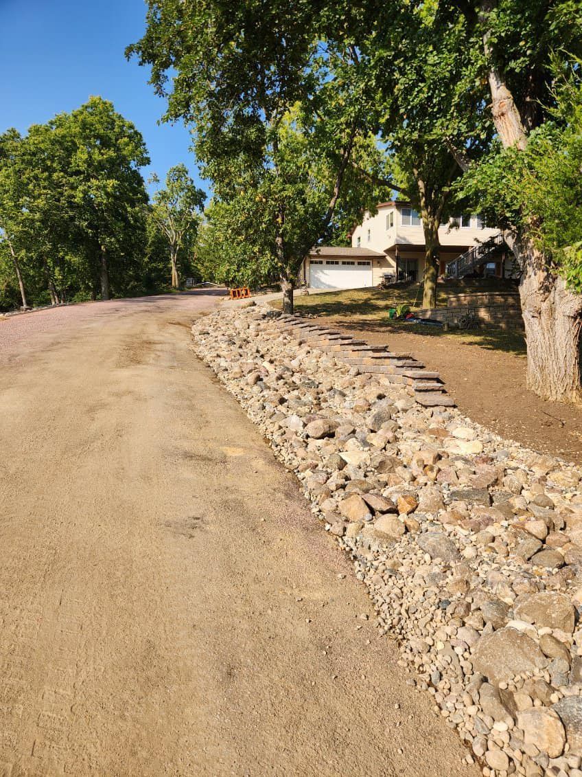 A stone retaining wall bordering a dirt driveway in front of a house, surrounded by trees on a sunny day.