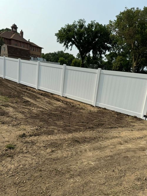 A long white vinyl privacy fence stands in a yard with dry, patchy grass, set against a background of trees and a house.