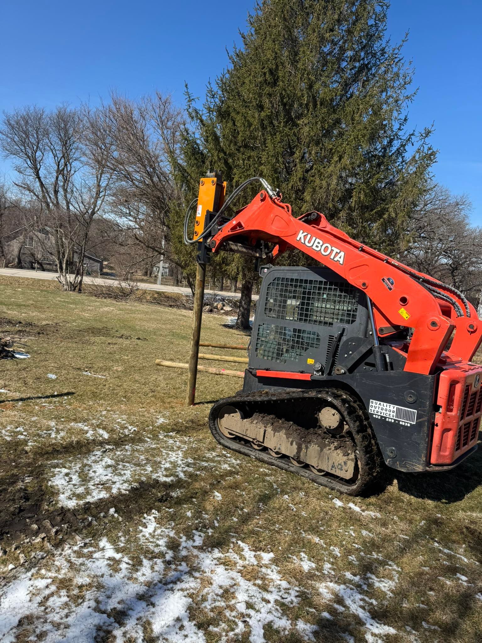 An orange Kubota track loader with a post driver attachment driving a wooden post into the ground in a grassy field.