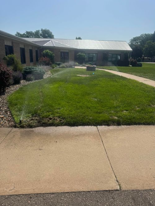 A brick building exterior with a green lawn being watered by an irrigation sprinkler system under a clear blue sky.