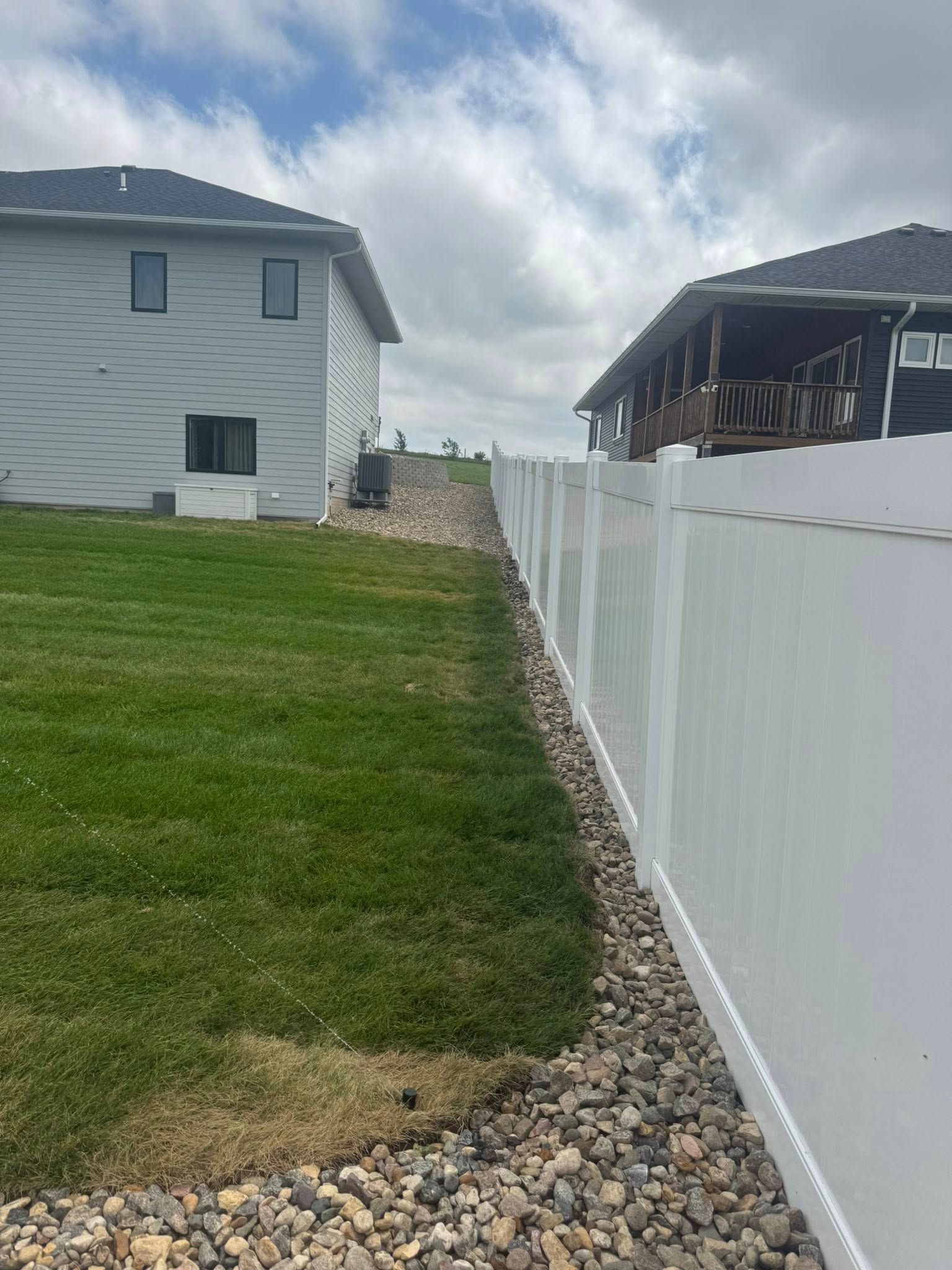 A side yard featuring a white vinyl fence bordering a green lawn and a narrow path of gray landscaping stones.