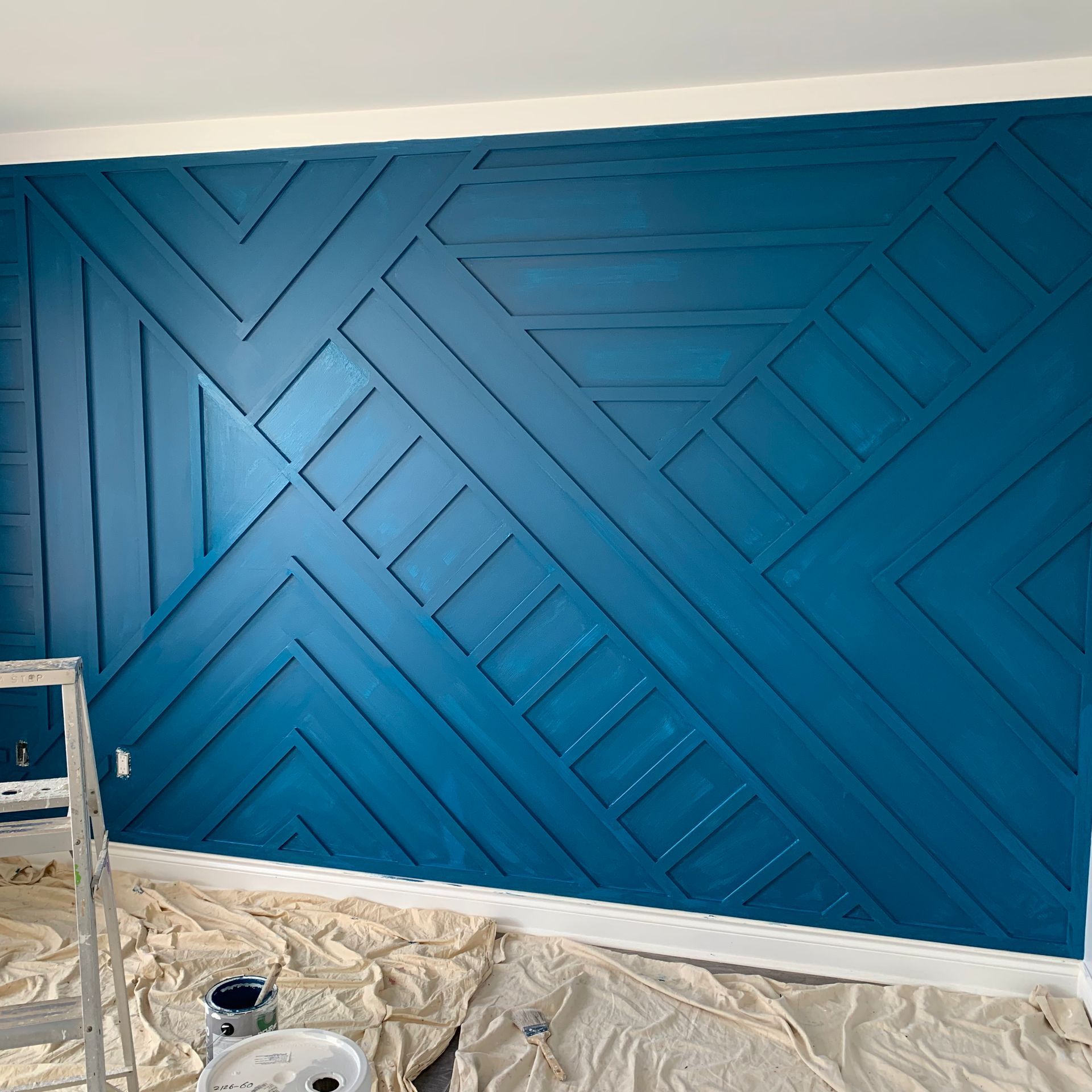Blue accent wall with geometric wood panel pattern, partially painted, with drop cloths and a ladder below