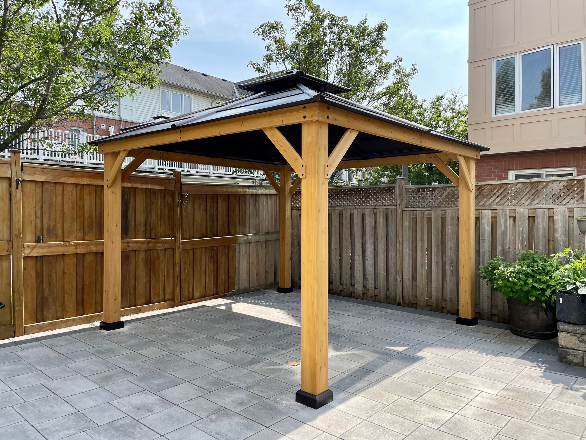 Wooden gazebo with a dark roof on a gray stone patio in a fenced backyard