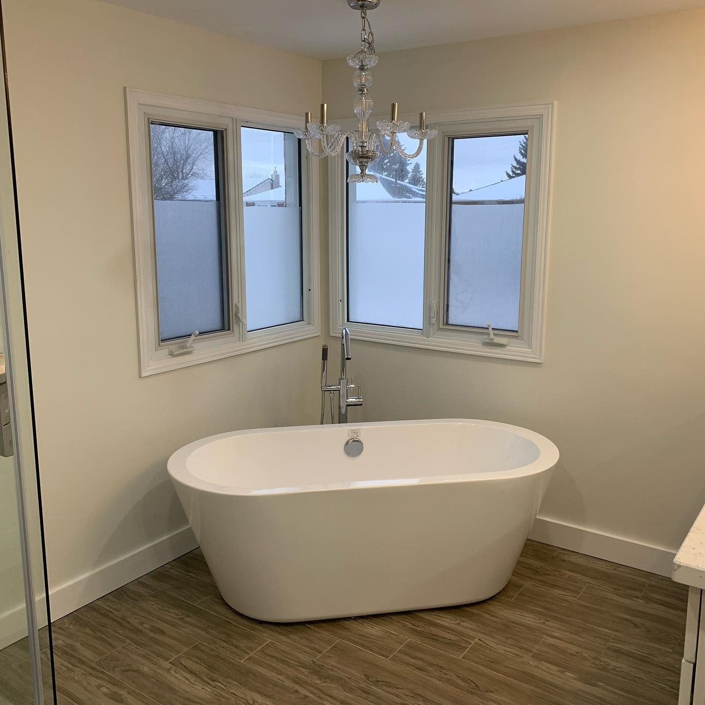 Modern bathroom with freestanding oval tub under corner windows and chandelier lighting