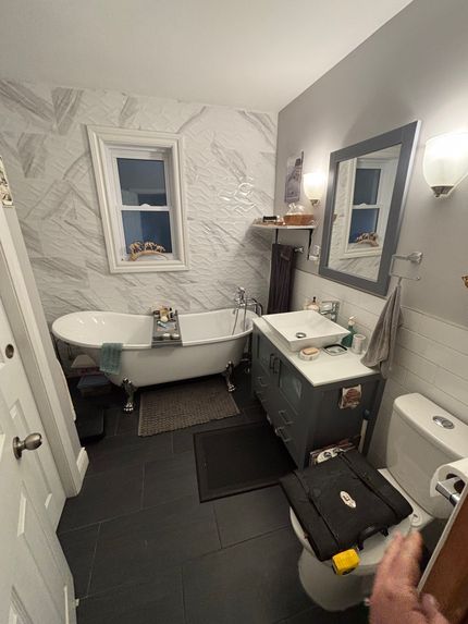 Small bathroom with white tub, toilet, sink vanity, gray walls, and cleaning supplies on the floor