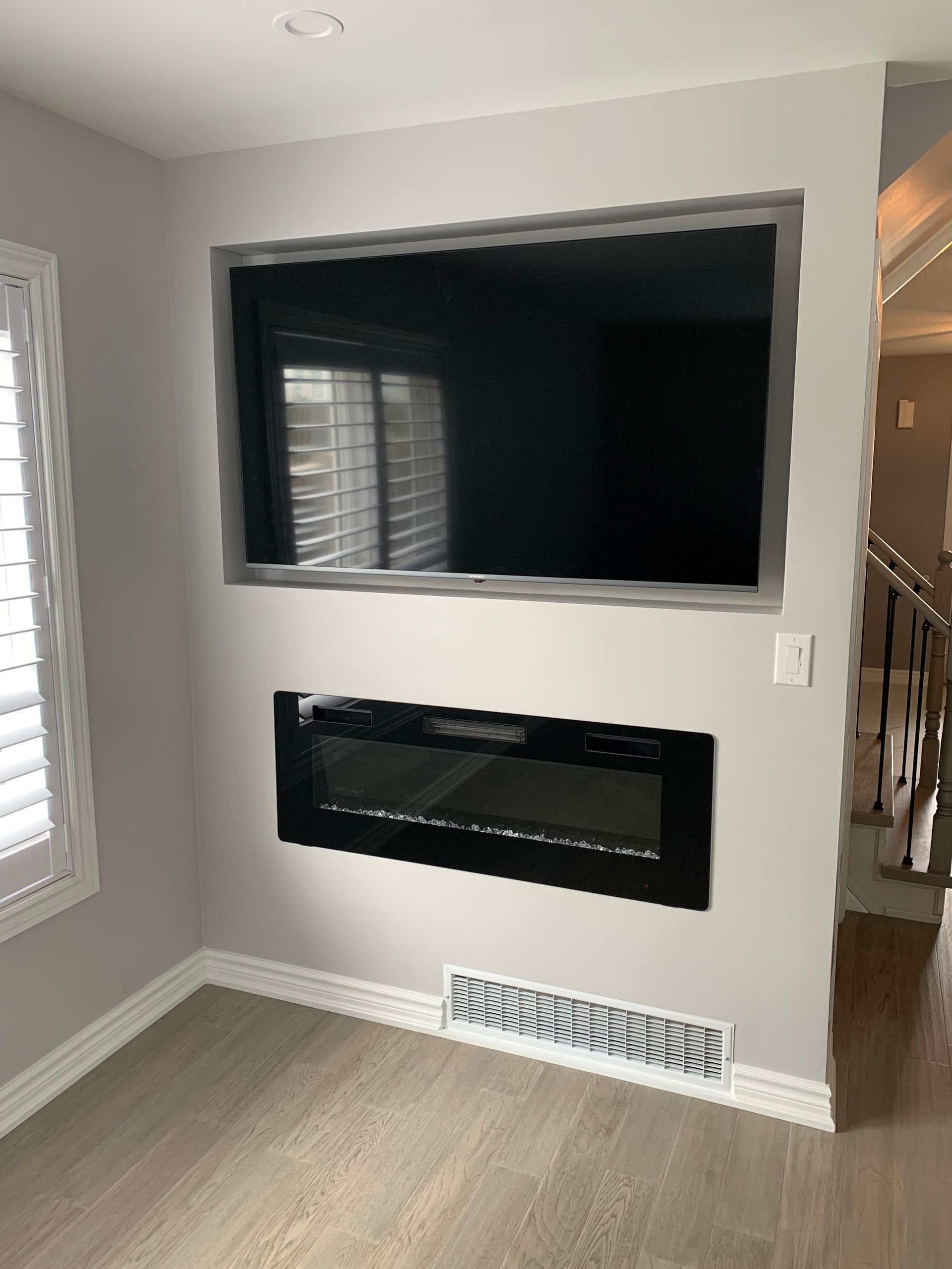 Living room wall with large TV, electric fireplace, and white vent below a window beside stairs