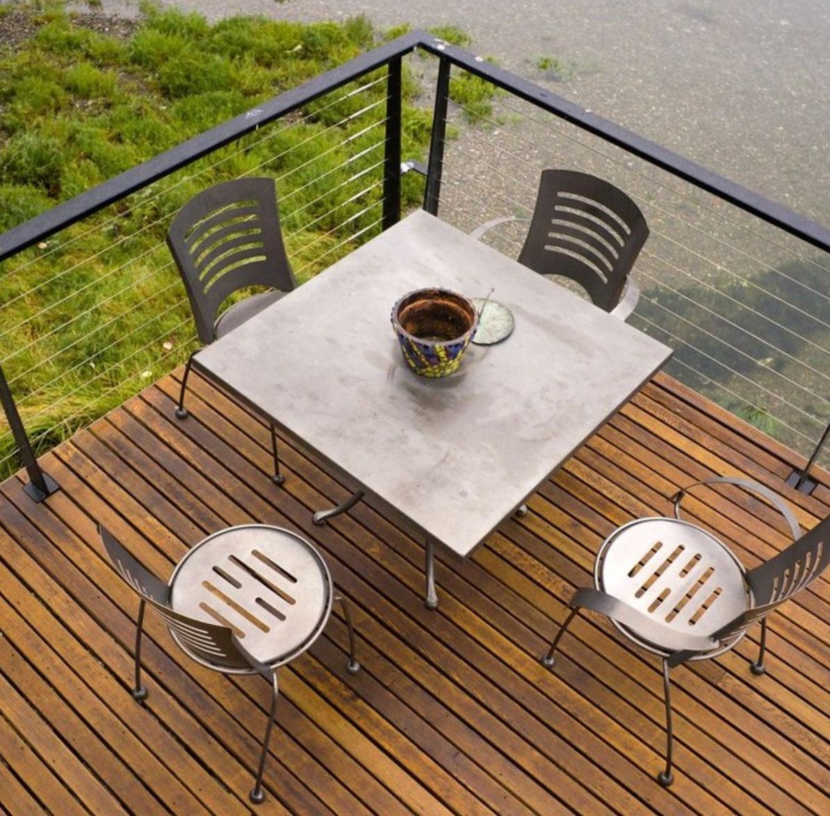 Small patio with a square table, four chairs, and a mug on a wooden deck by a glass railing.