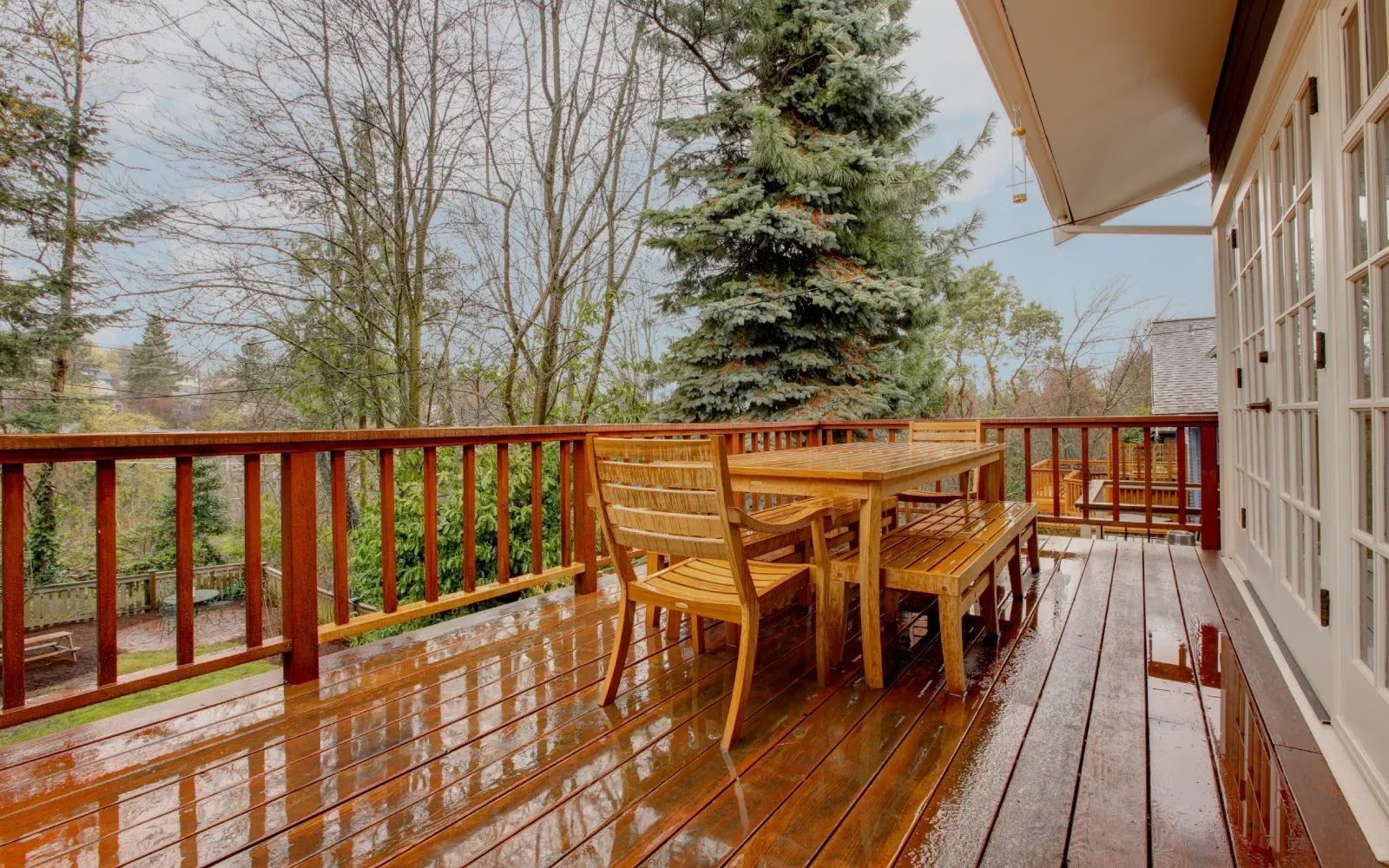 Wooden deck with wet railing, wicker table and chairs, overlooking trees beside a house
