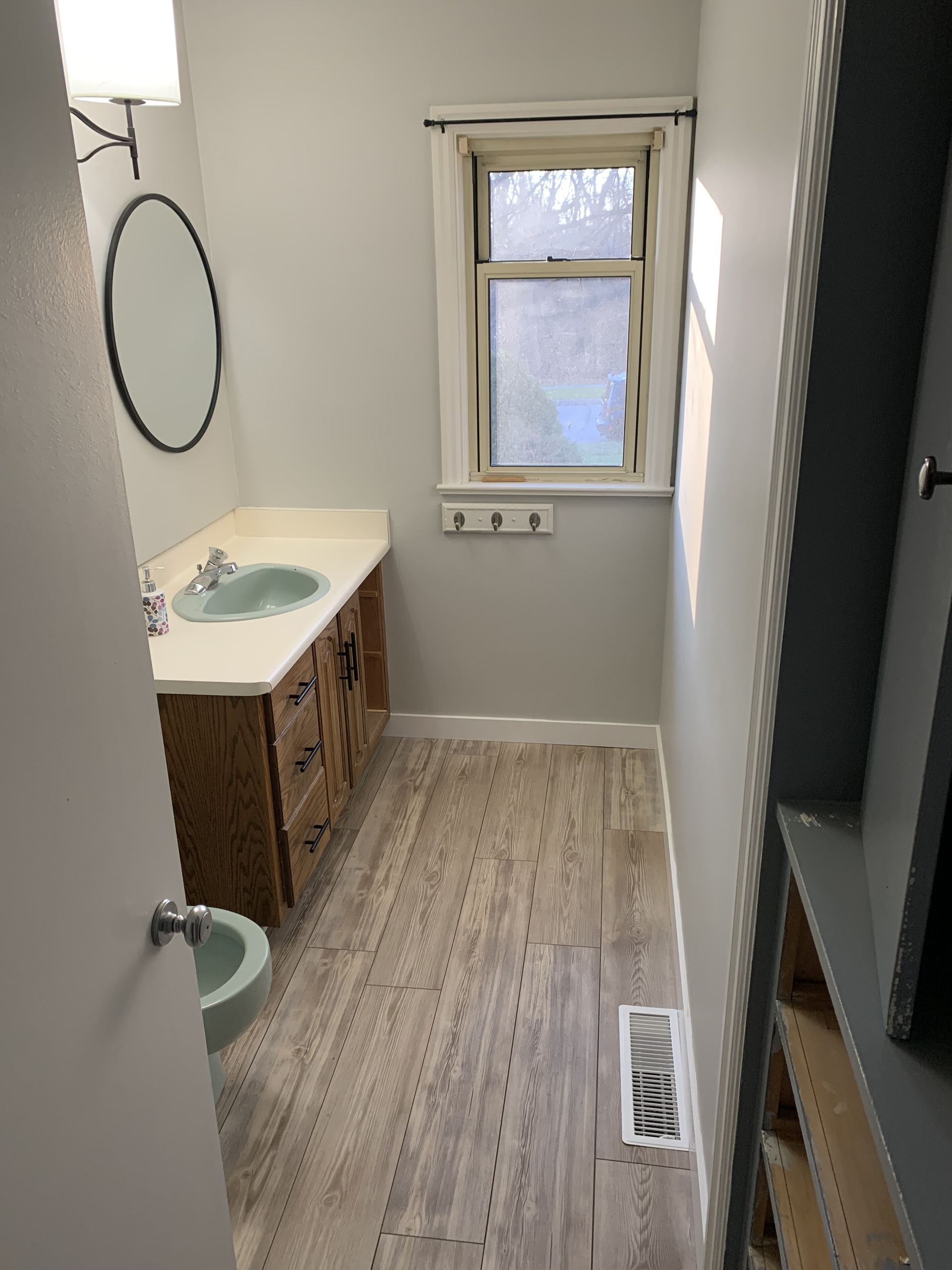 Narrow bathroom with vanity, toilet, window, and wood-look floor.