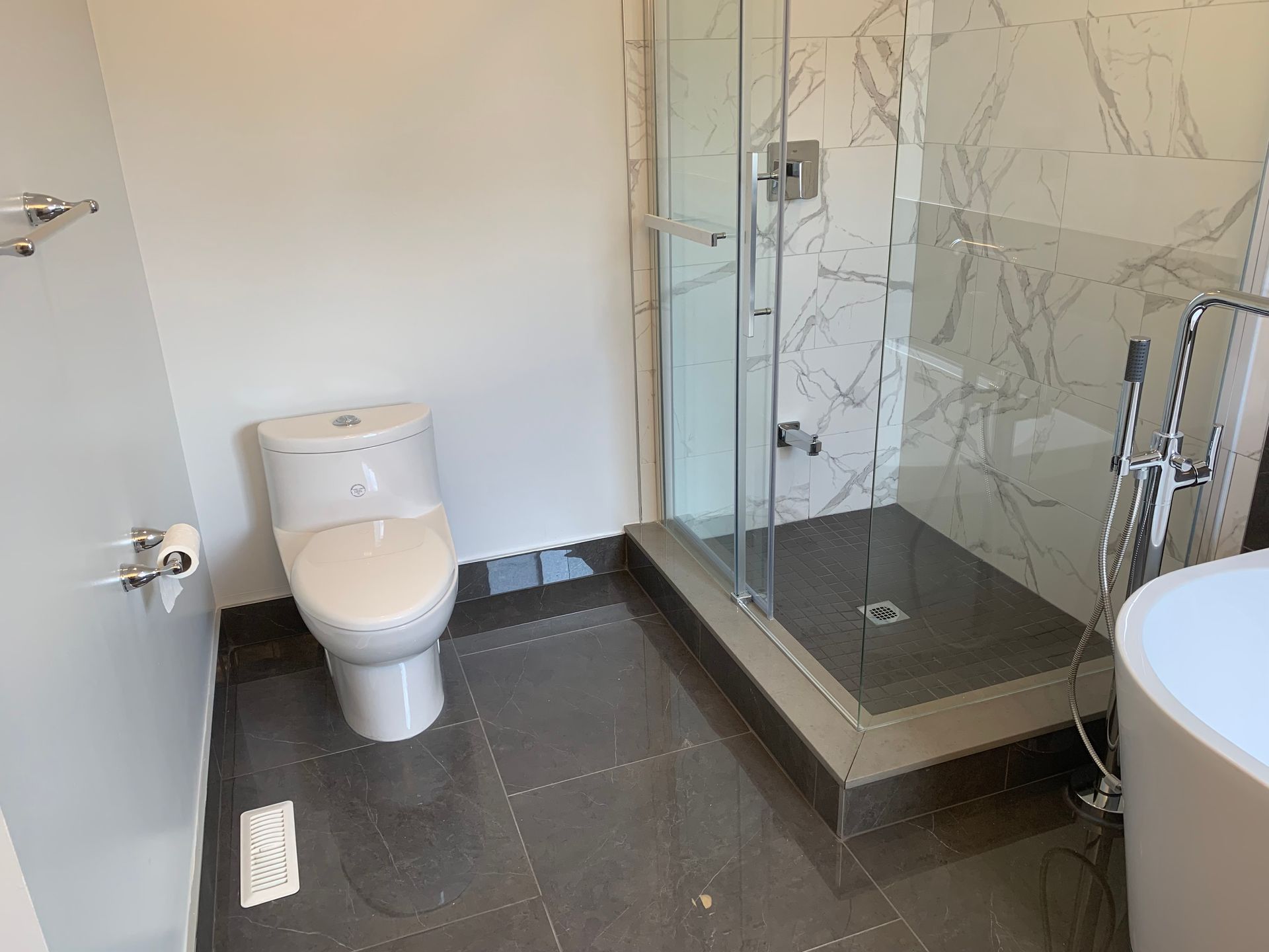 Small modern bathroom with toilet, glass shower, dark tile floor, and sink partially visible.