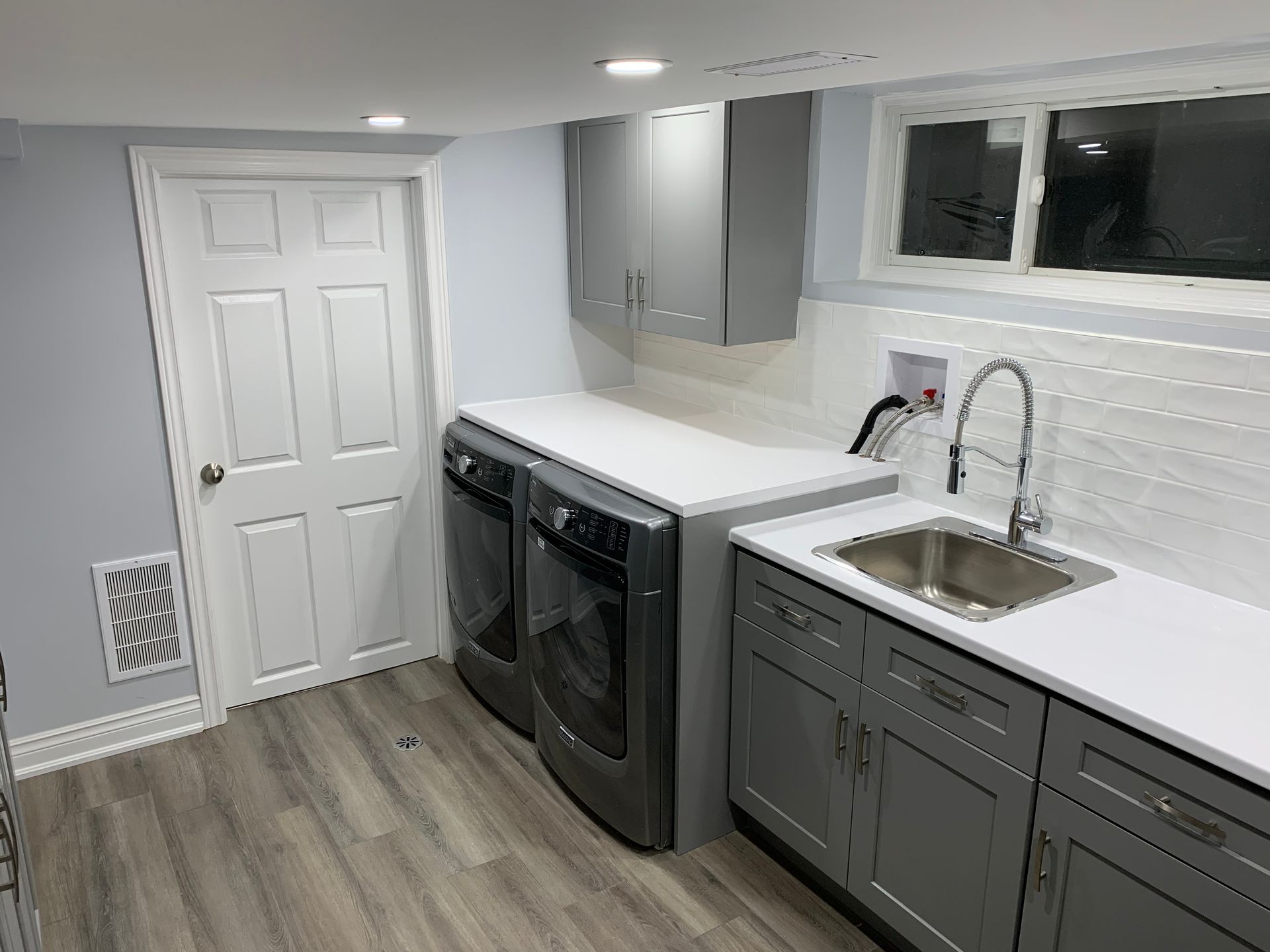 Bright laundry room with gray cabinets, white counters, sink, washer and dryer, and wood-look floor.