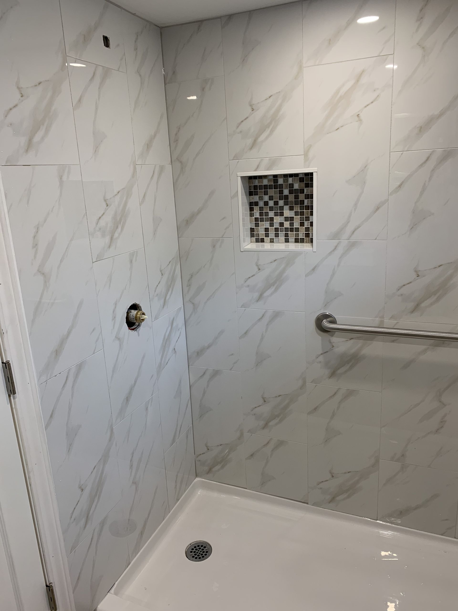 White tiled shower stall with built-in shelf, grab bar, and drain on the floor