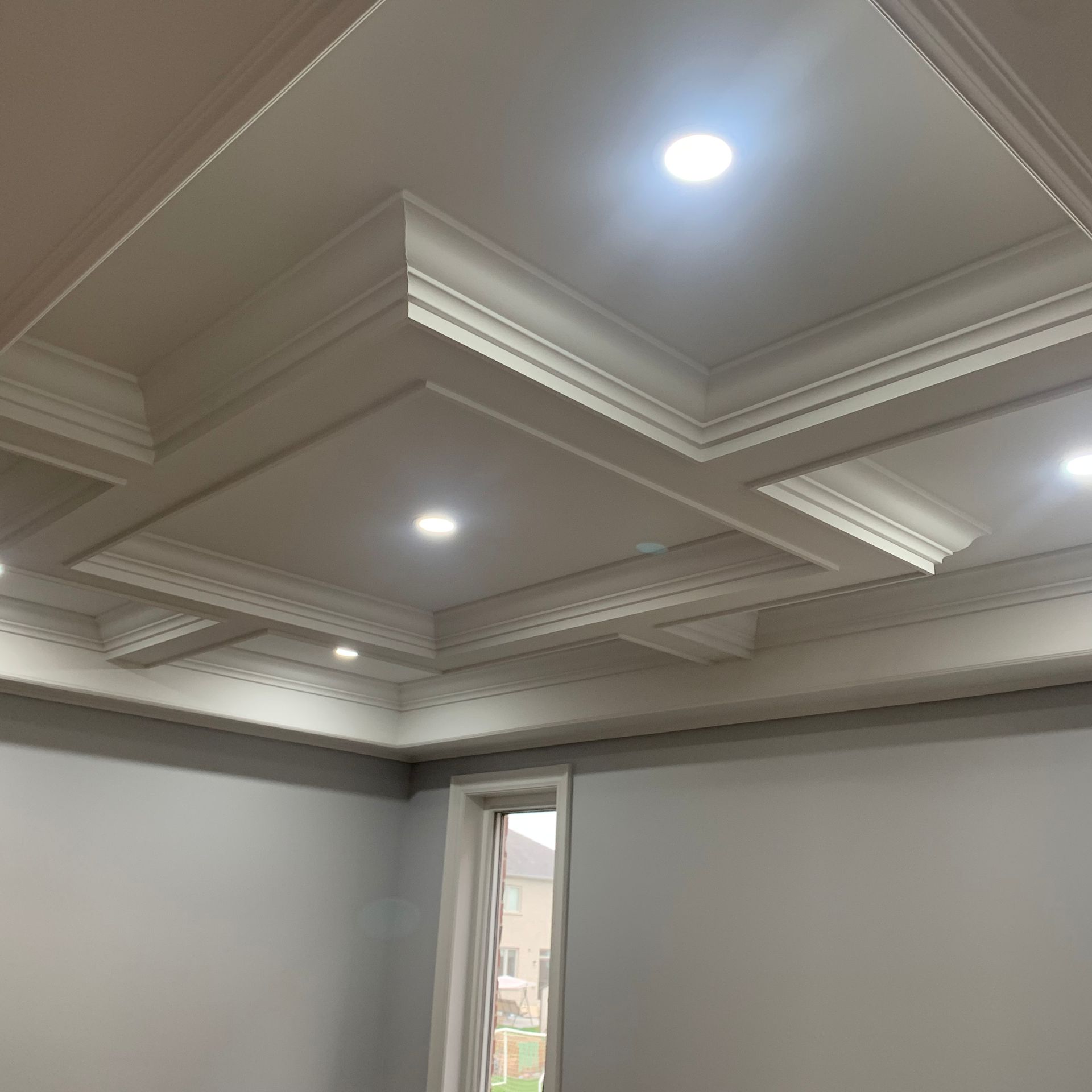 Empty room with ornate white coffered ceiling and recessed lights, viewed from a corner.