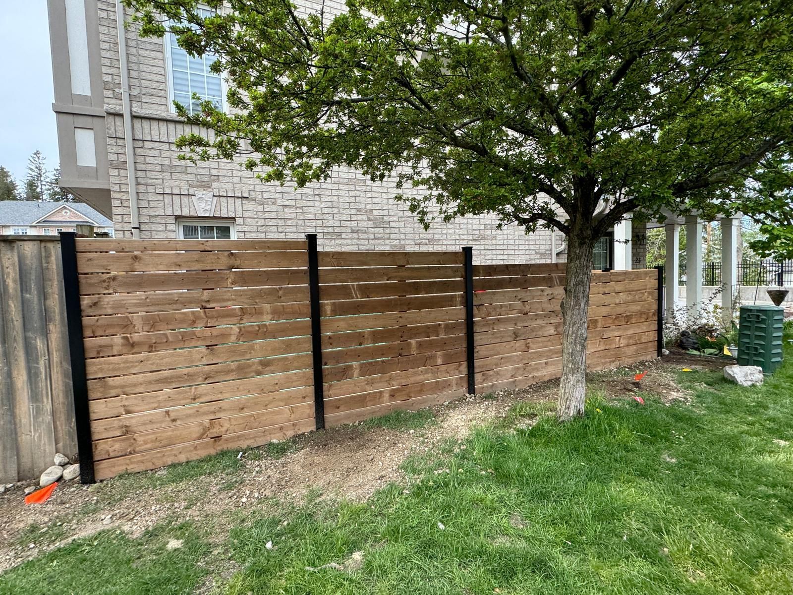 Wooden privacy fence with black posts beside a grassy yard and tree near a stone building