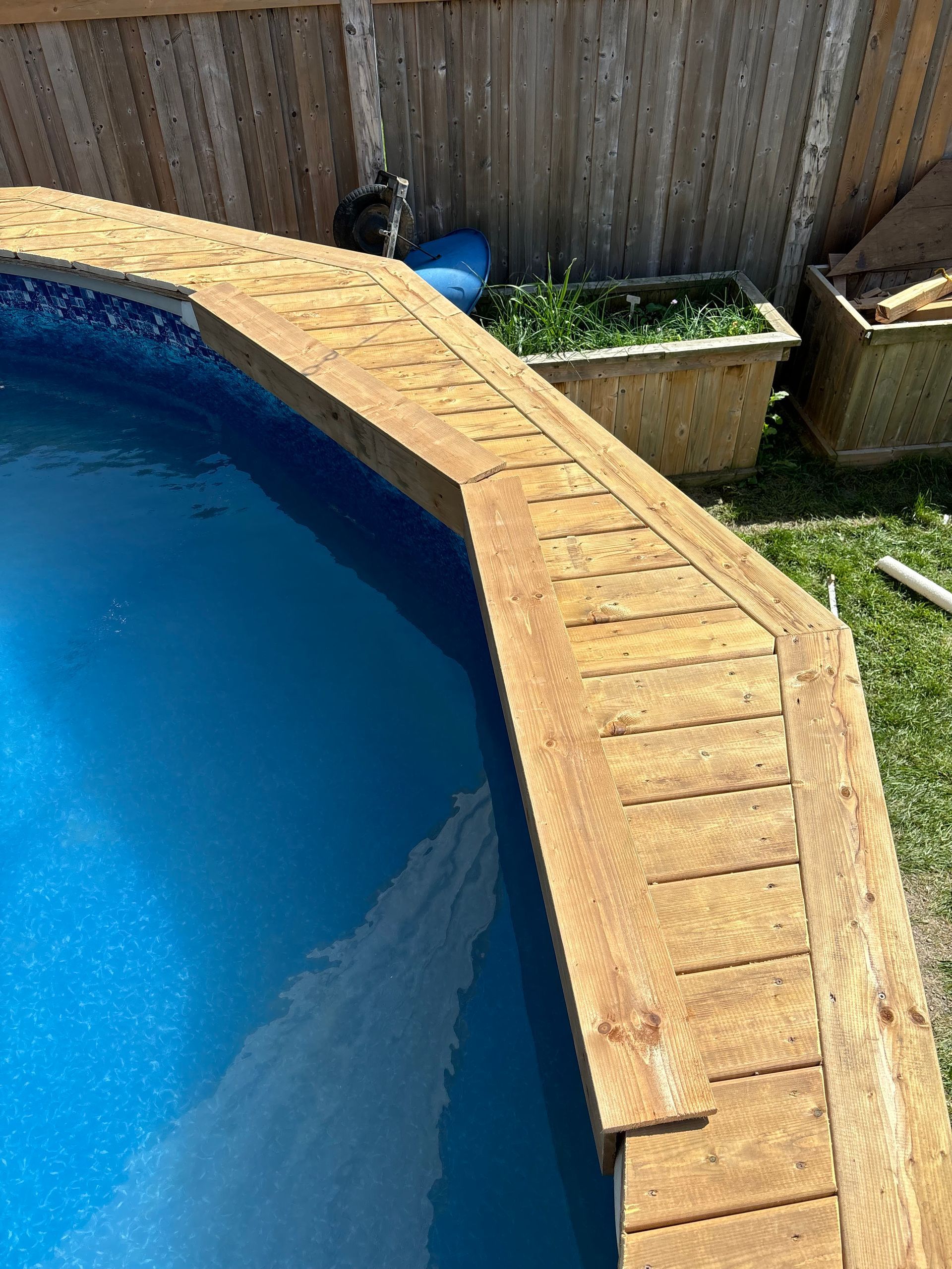 Curved above-ground pool with blue water and wooden deck edging beside a fenced yard