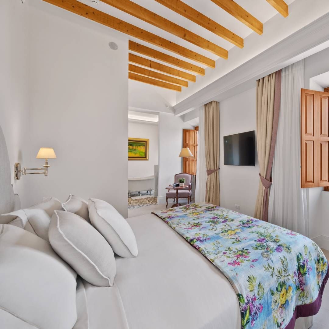 Bedroom with white walls, wooden beams, a bed with floral cover, and a window with shutters.
