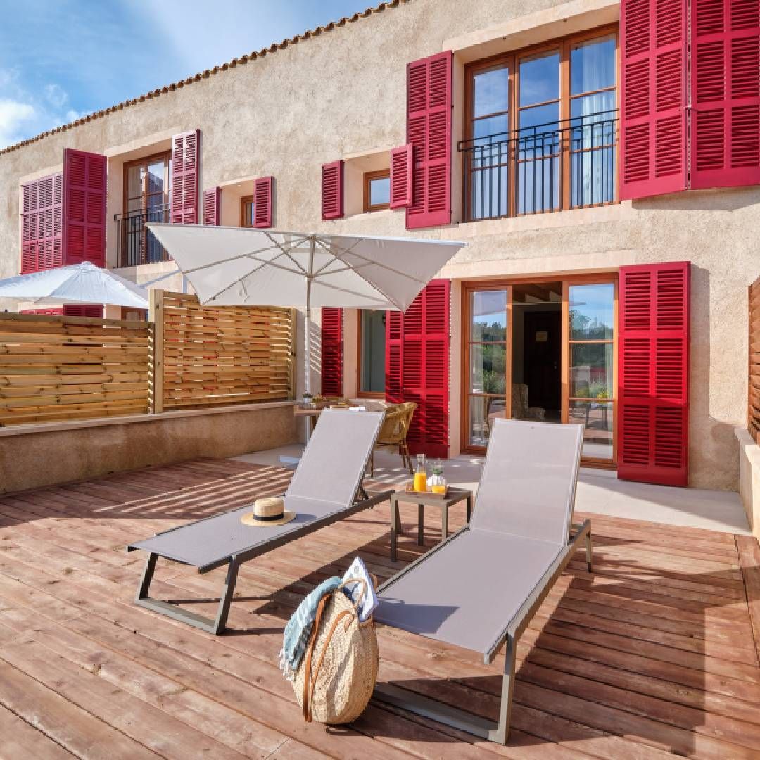 Two lounge chairs under an umbrella on a wooden deck in front of a building with red shutters.