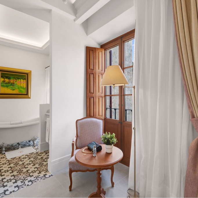 Cozy corner of a room with a small table, chair, and a window with wooden shutters. A lamp stands nearby.