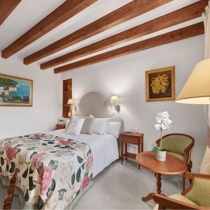 Bedroom with floral bedspread, wooden beams, artwork, and seating.