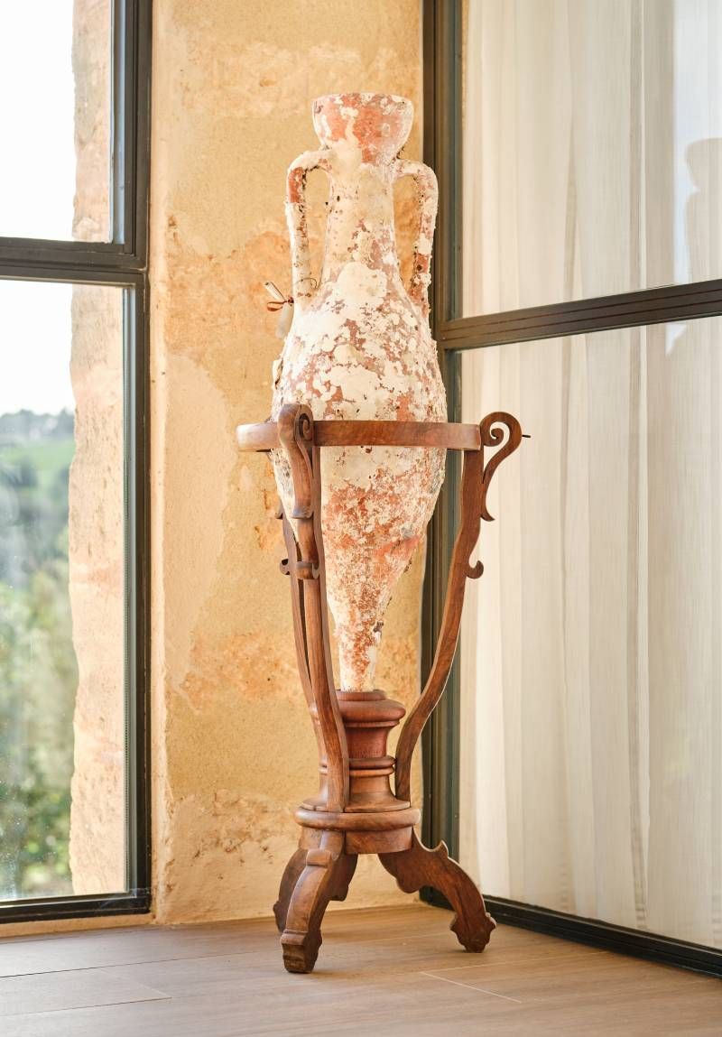 An antique vase on a wooden stand in front of a window. The vase has an aged, worn appearance.