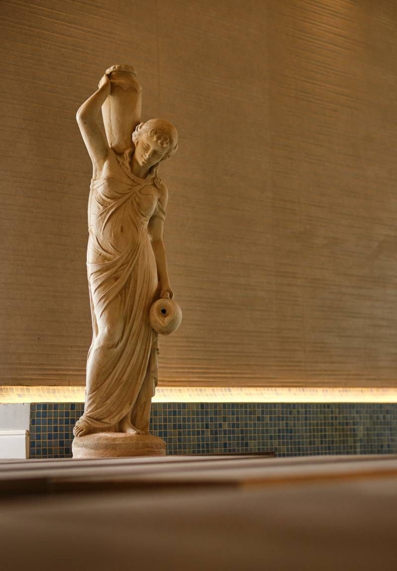 Statue of a person carrying water jars, standing on a tiled surface, with textured wooden paneling in the background.