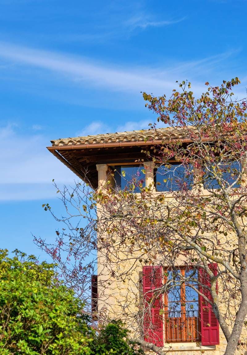 Stone building with red shutters, under blue sky, tree with reddish leaves in foreground.