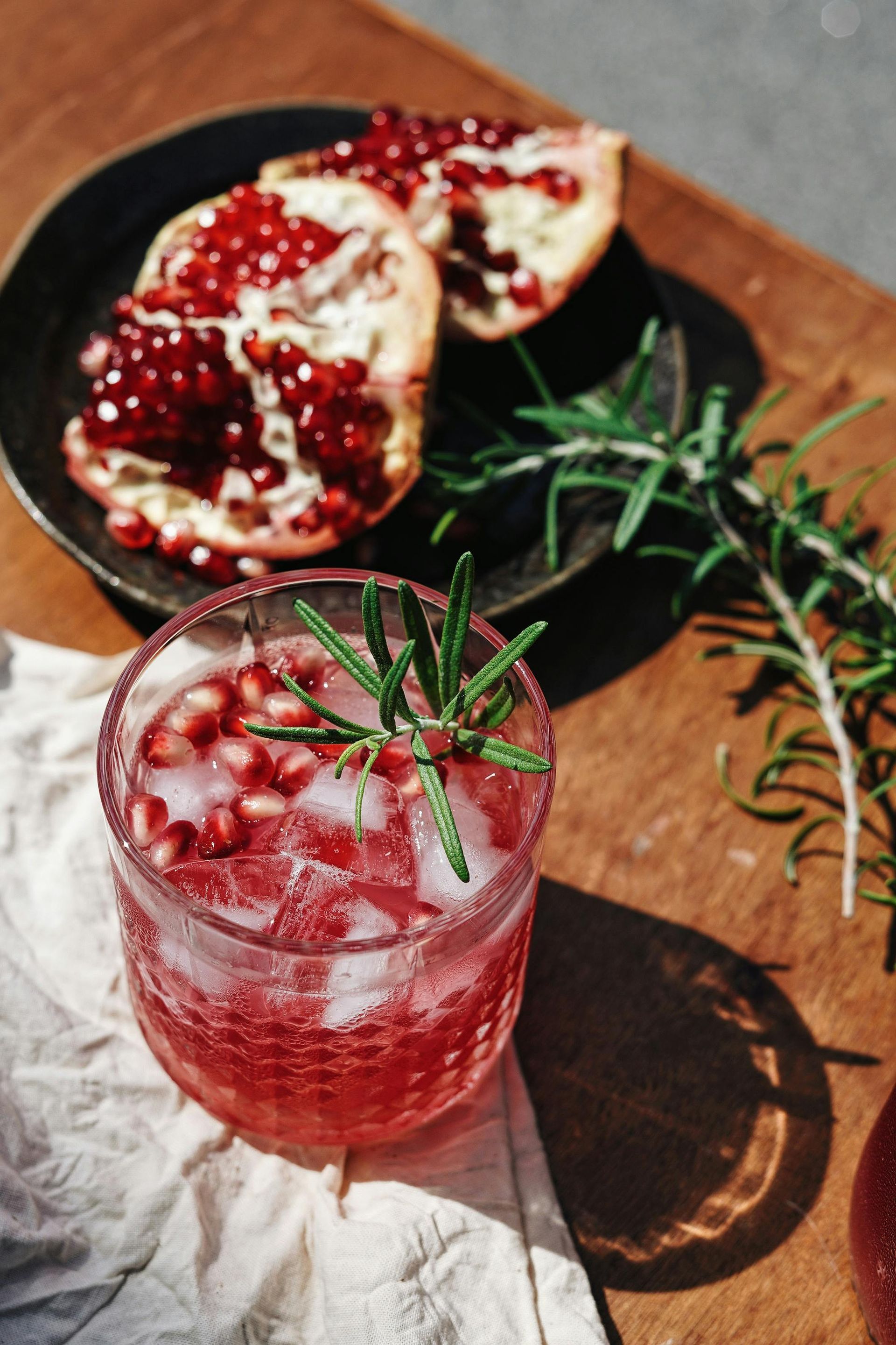 Pomegranate cocktail with rosemary garnish and pomegranate seeds on a wooden table.