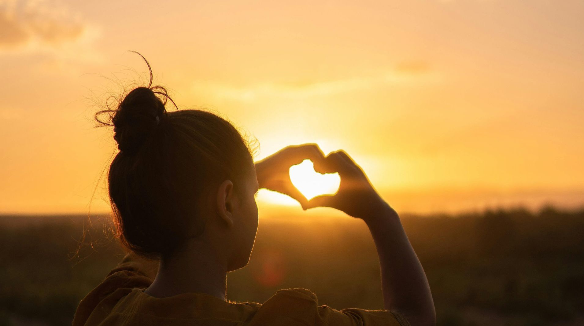 A woman is making a heart shape with her hands in front of the sun at sunset.