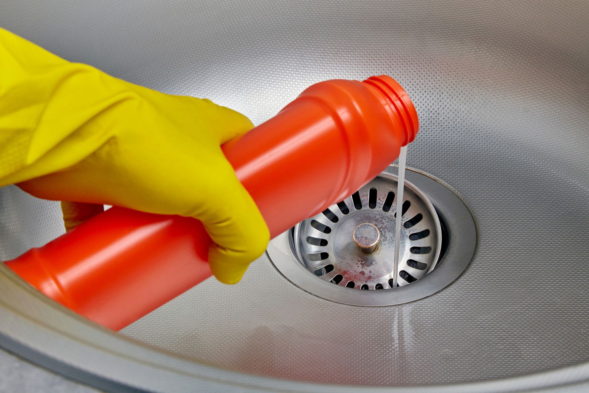 Gloved hand pouring drain cleaner from a red bottle into a stainless steel sink.