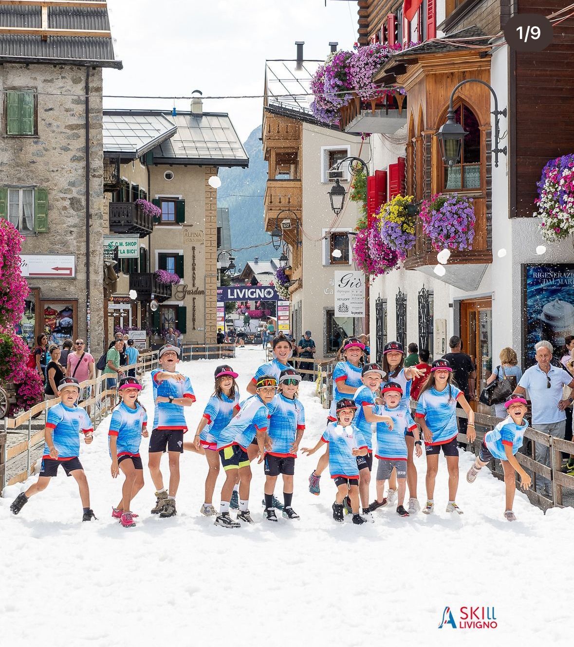 Bambini con magliette blu posano su una strada innevata, davanti a edifici con fioriere.