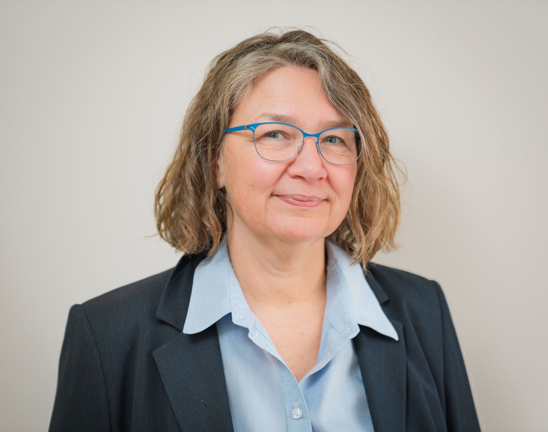 Woman with glasses and short brown hair, wearing a blue shirt and navy blazer, smiling at the camera.
