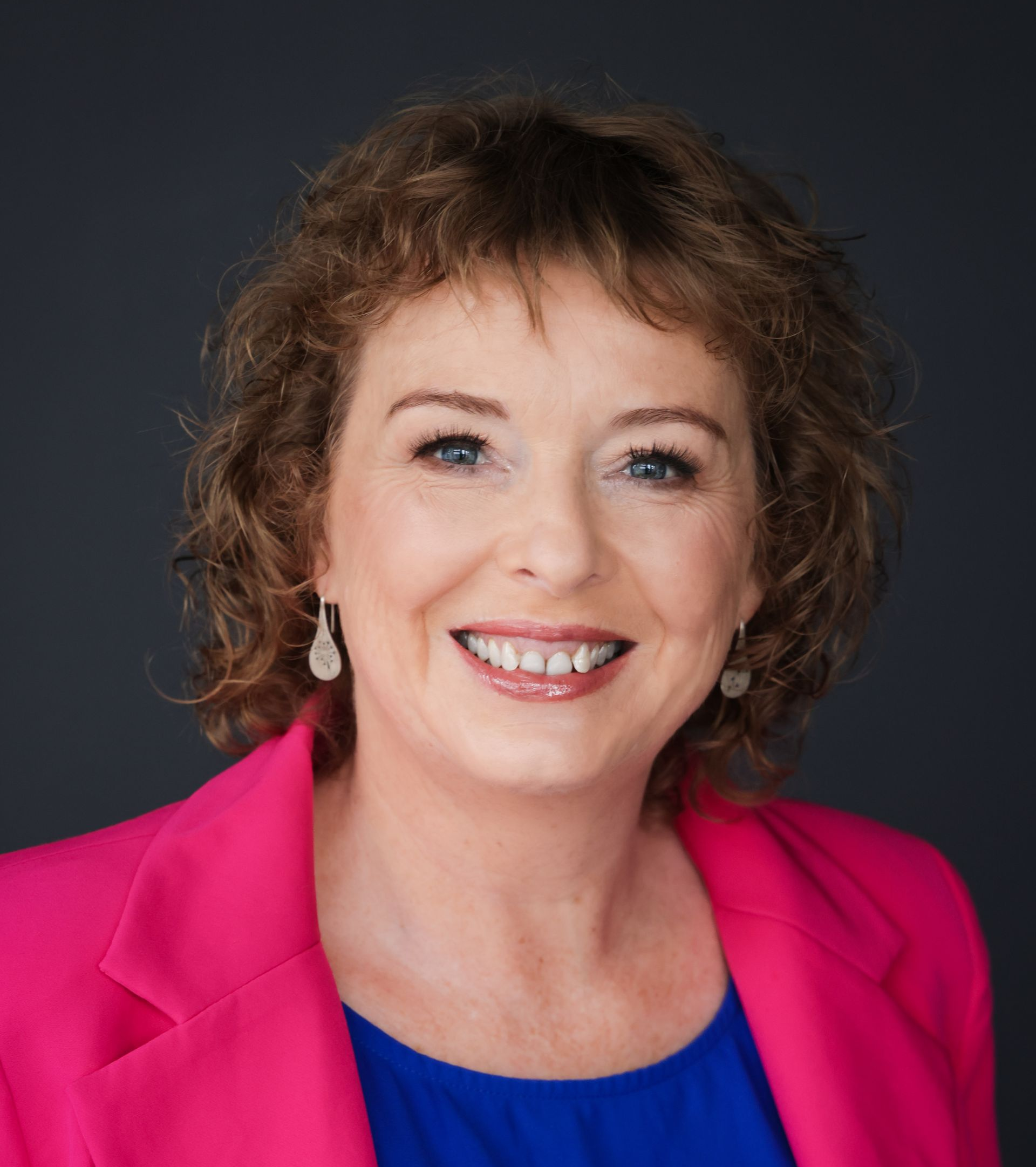 Woman smiling, wearing a pink blazer over a blue top, with shoulder-length curly hair; dark gray background.