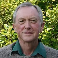 Man smiling, wearing a green collared shirt and gray sweater. Outdoors with leafy green background.